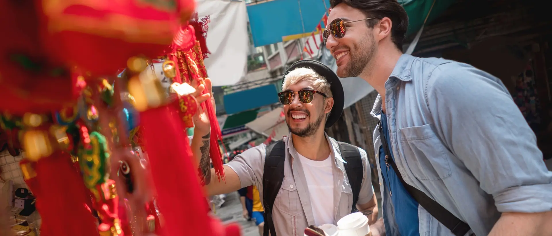 Two smiling men explore a vibrant market, admiring colorful decorations while holding a coffee cup and a guide.