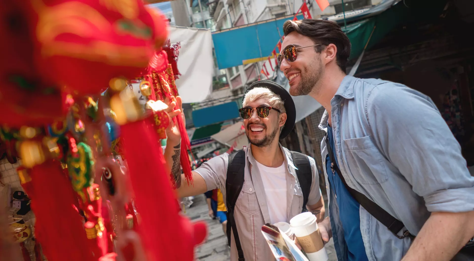 Two smiling men explore a vibrant market, admiring colorful decorations while holding a coffee cup and a guide.