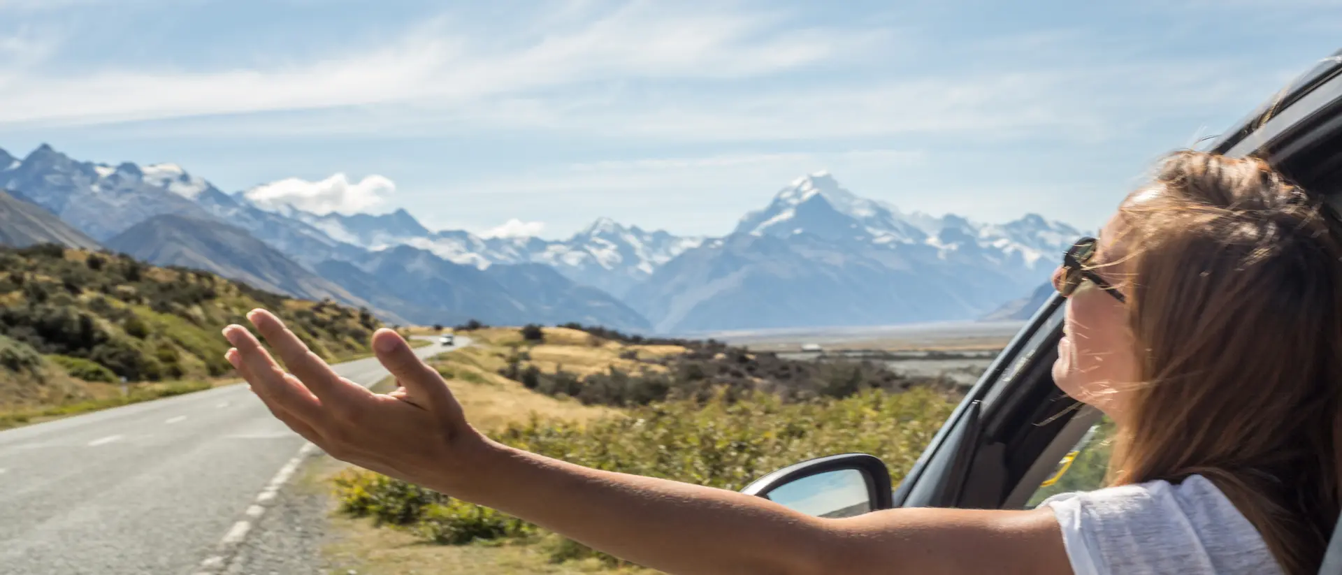 A person leans out of a car window, enjoying a scenic view of mountains and open road on a sunny day.