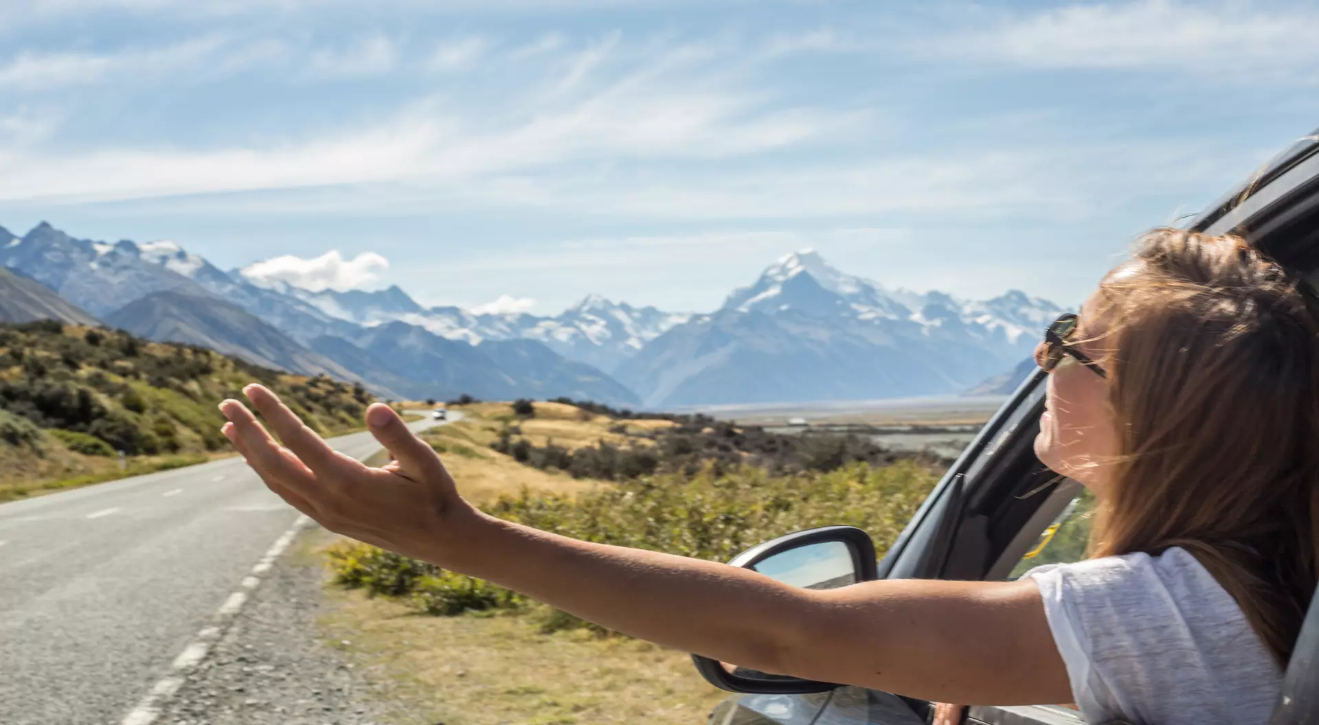 A person leans out of a car window, enjoying a scenic view of mountains and open road on a sunny day.