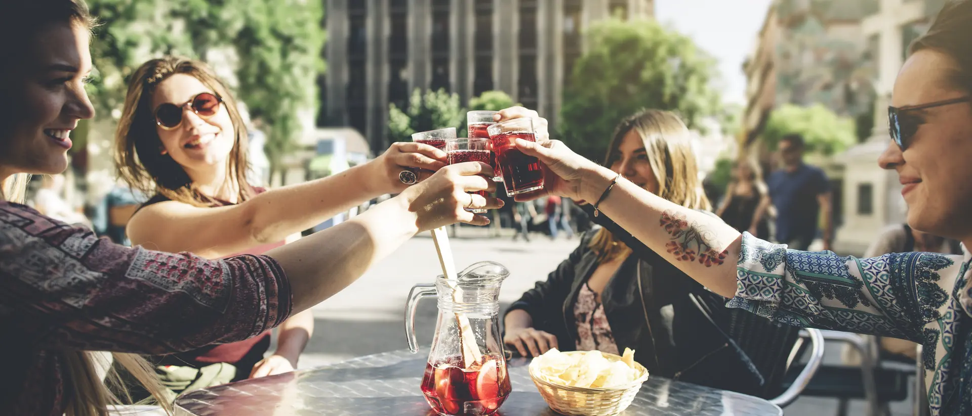 Four friends clink glasses of drinks at an outdoor table, enjoying