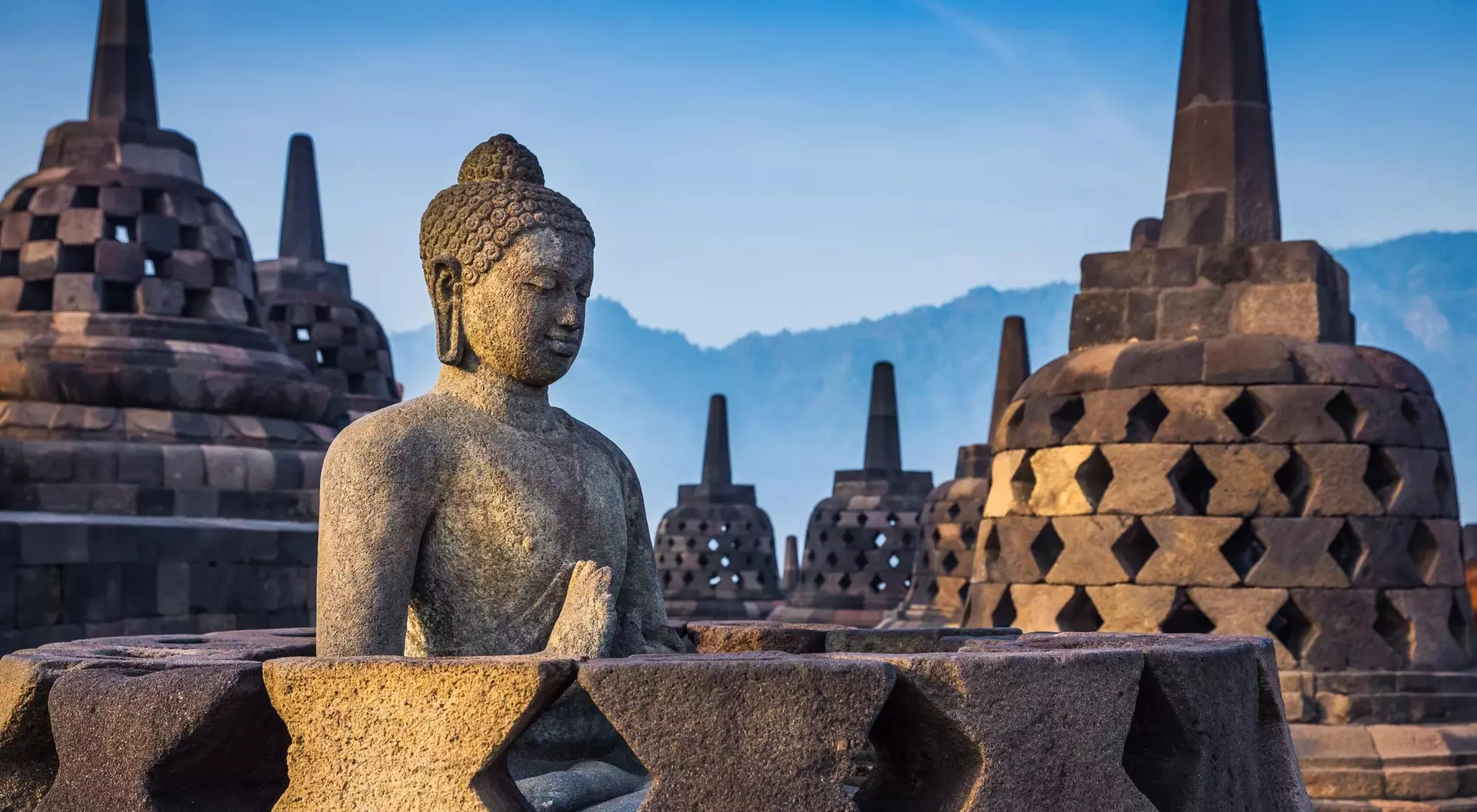 Buddha statue in the foreground with ancient, tiered stupas against a hazy mountain backdrop at sunrise.