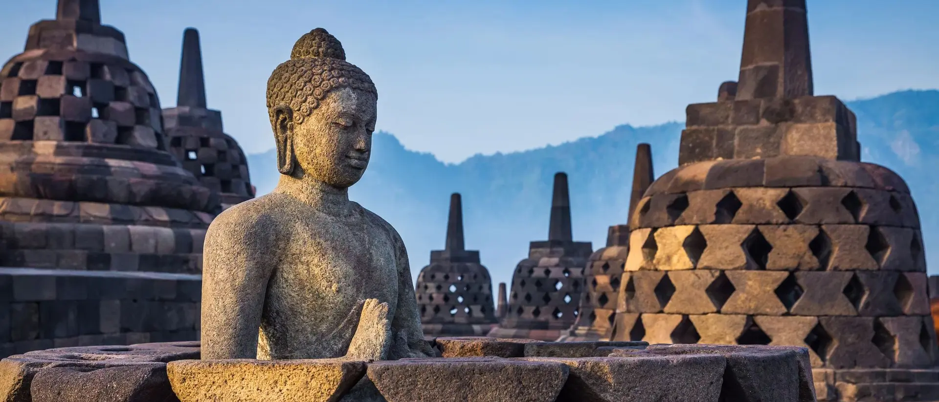 Buddha statue in the foreground with ancient, tiered stupas against a hazy mountain backdrop at sunrise.