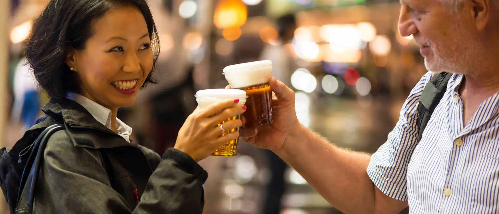 A woman and a man cheerfully clink their beer glasses together in a lively urban setting at night.