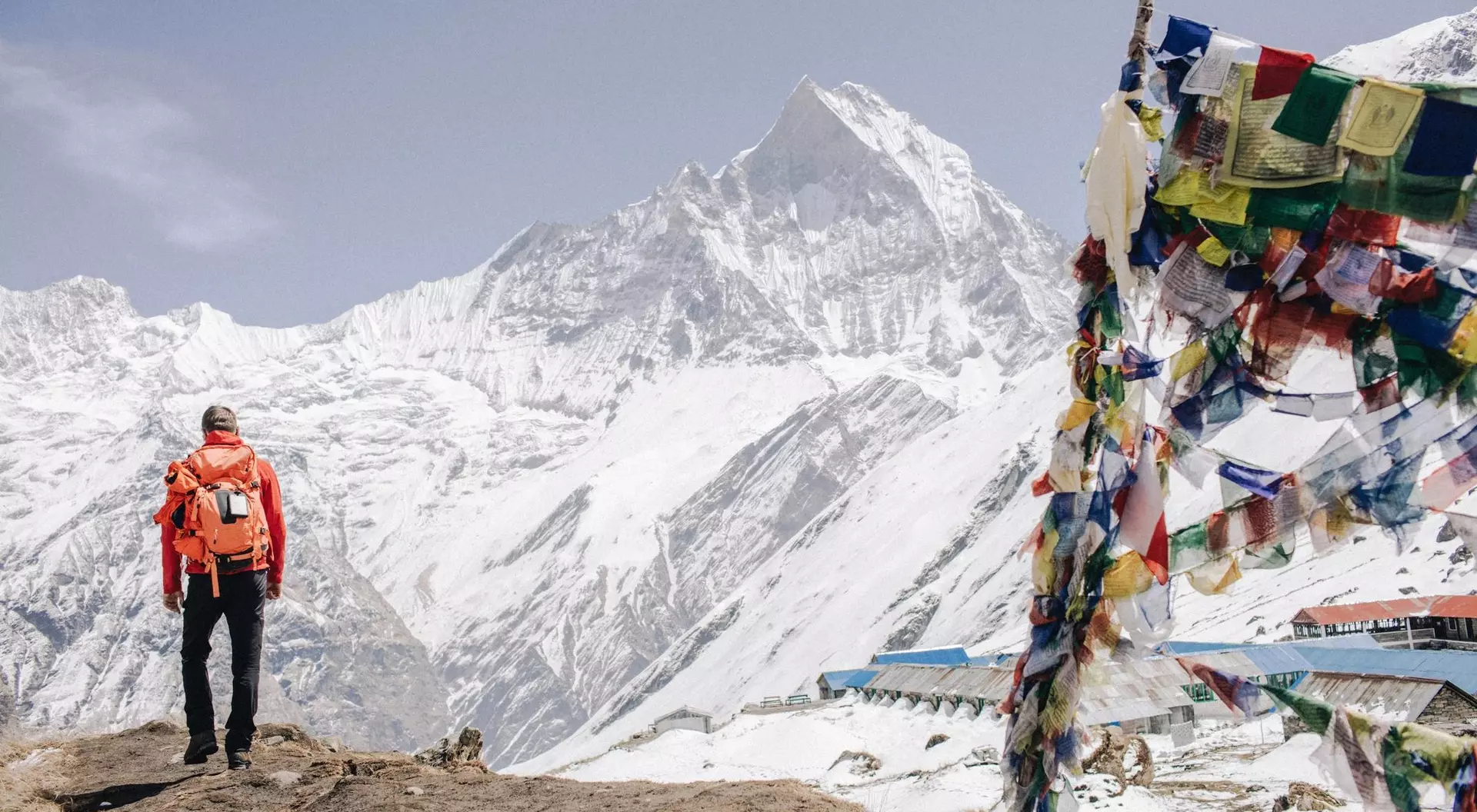 Hiker in an orange jacket stands on a rocky outcrop, surrounded by snow-capped mountains and colorful prayer flags.