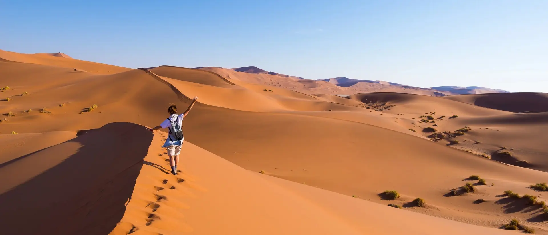 A person walks on a sandy dune in a desert, with rolling hills and a clear blue sky in the background.