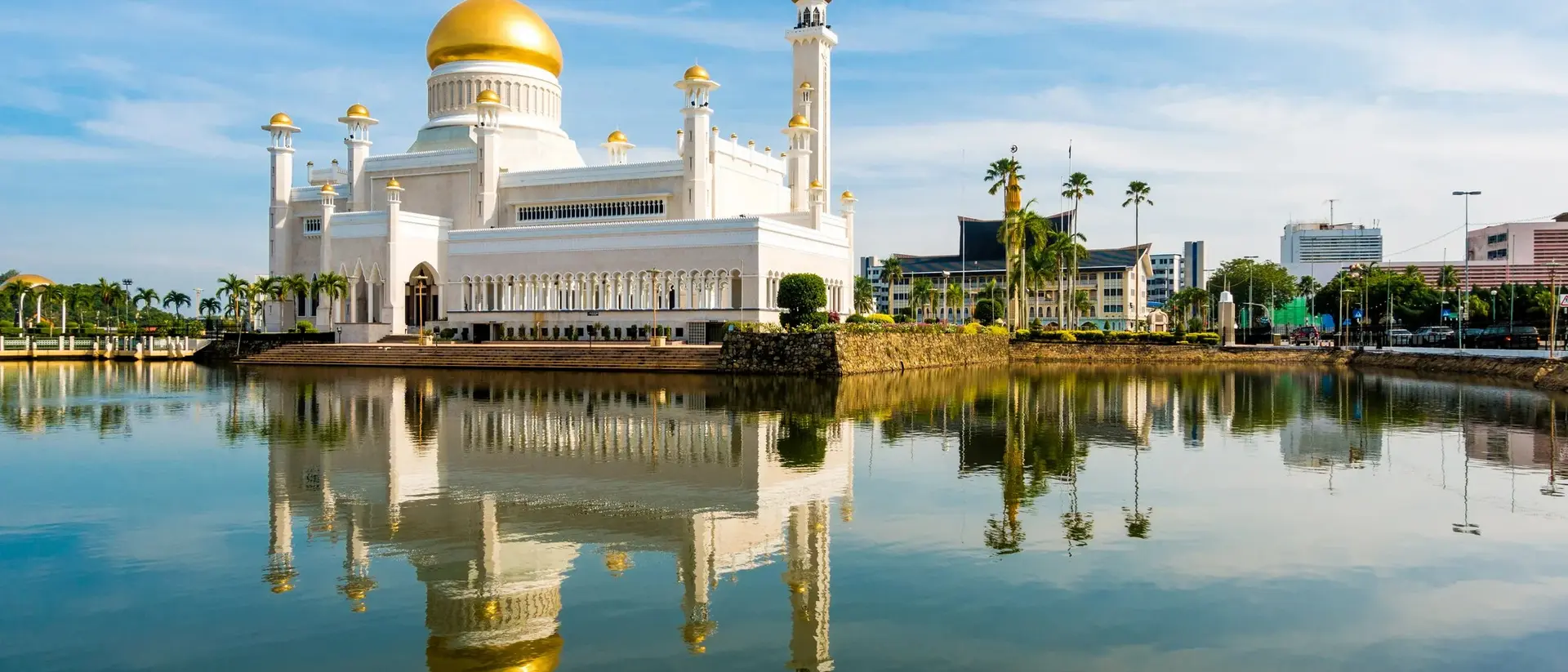 A grand white mosque with a gold dome reflects in a tranquil lake, under a blue sky with scattered clouds.