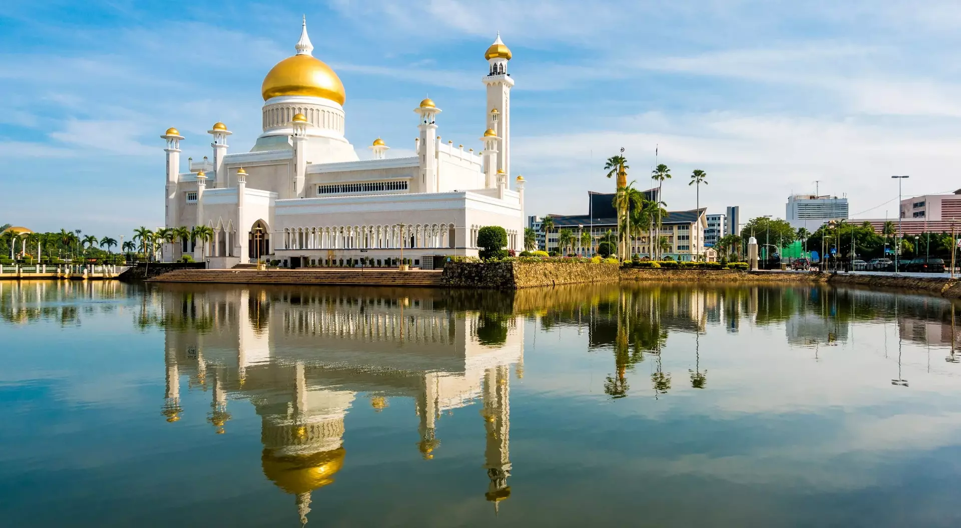 A grand white mosque with a gold dome reflects in a tranquil lake, under a blue sky with scattered clouds.