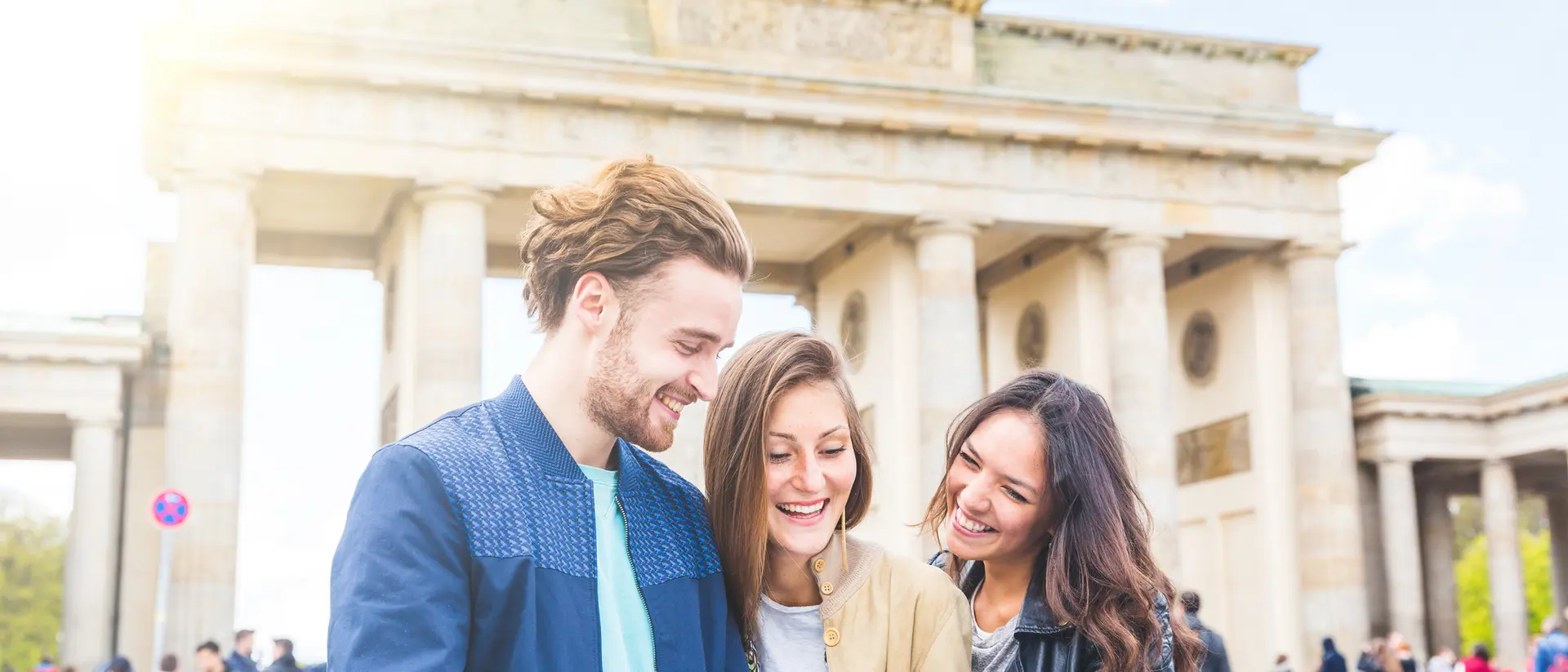 Three friends smiling and examining a map in front of the Brandenburg Gate, with people walking in the background.
