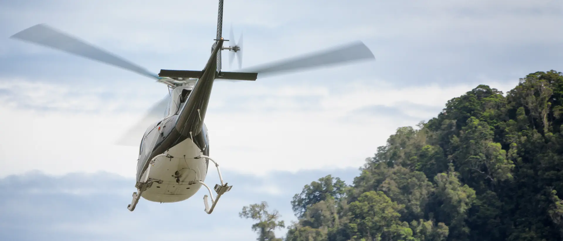 A helicopter flies above a lush green hillside under a cloudy sky.
