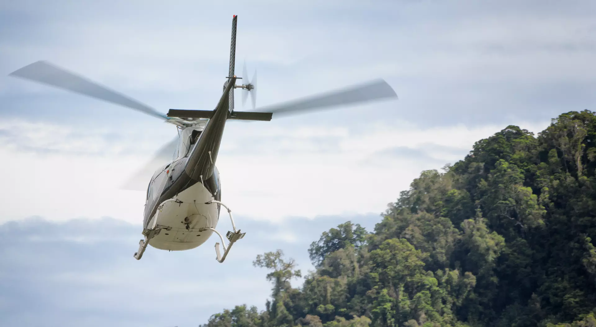 A helicopter flies above a lush green hillside under a cloudy sky.
