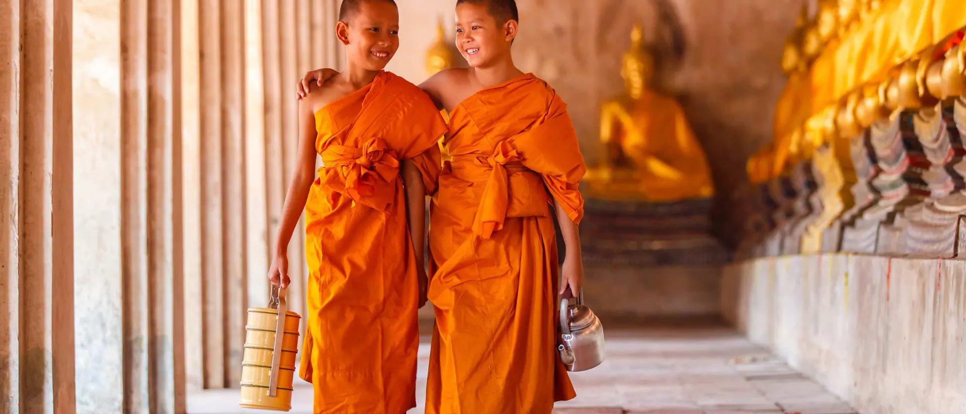 Two young monks in orange robes walking together, smiling, with golden statues in the background.