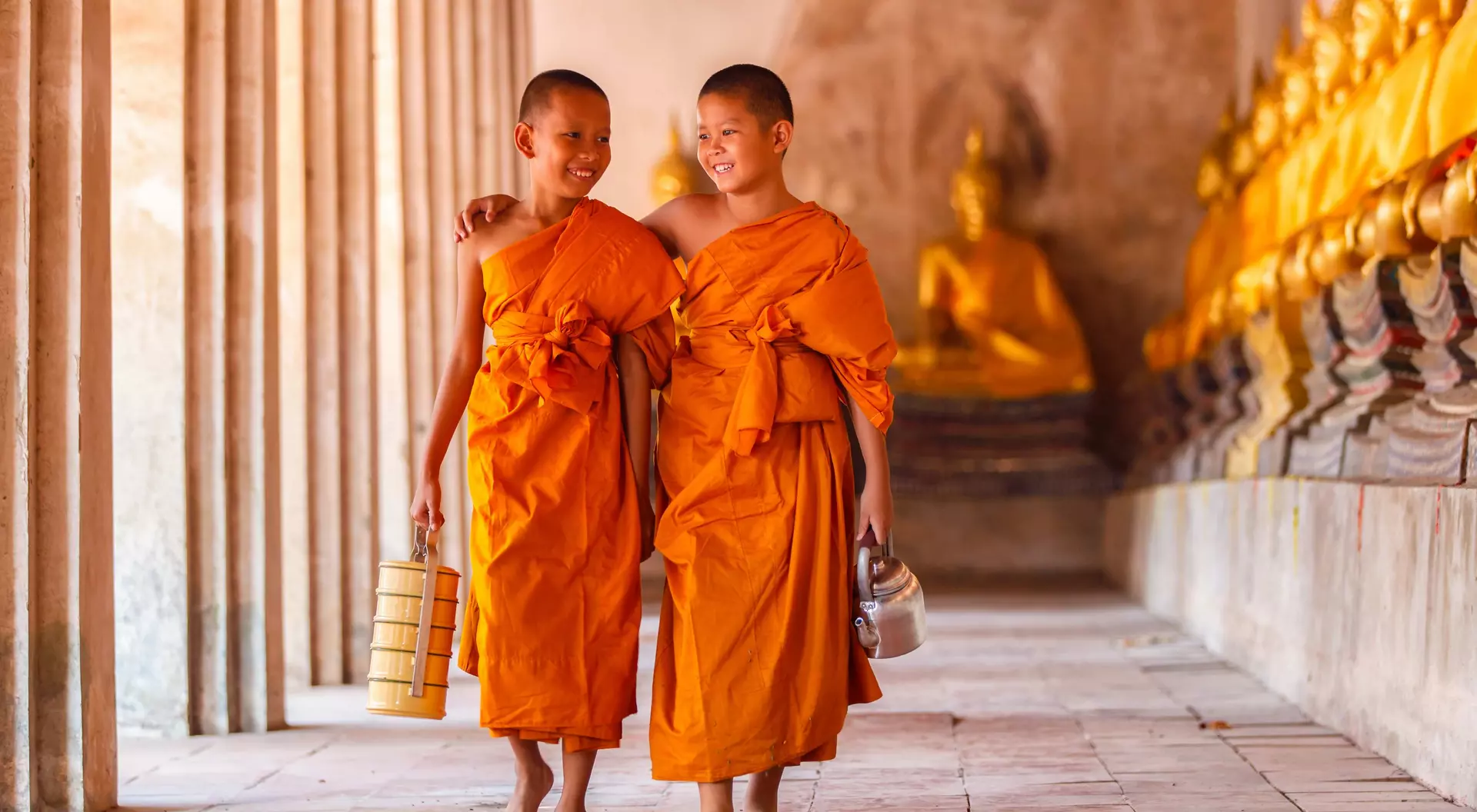 Two young monks in orange robes walking together, smiling, with golden statues in the background.