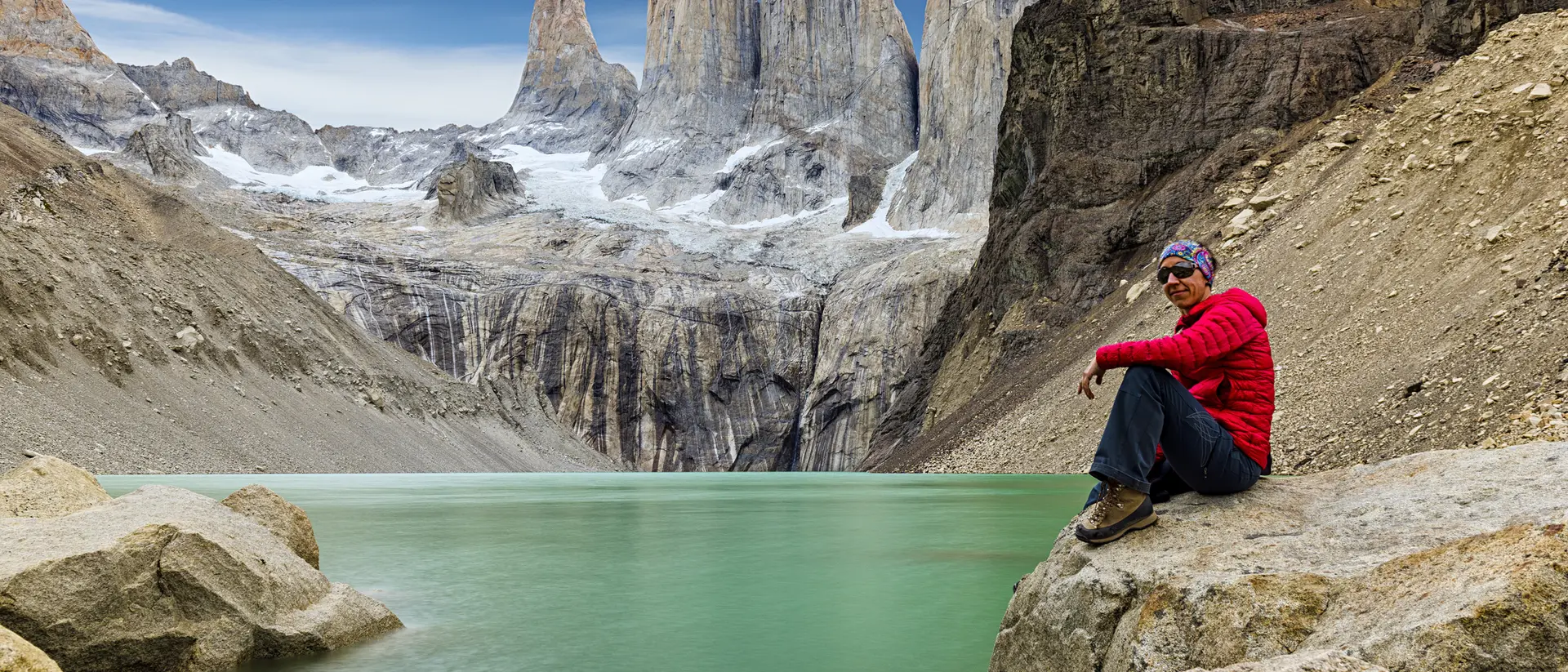 Person in red sitting on rocks by a turquoise lake, with towering mountains in the background under a clear blue sky.