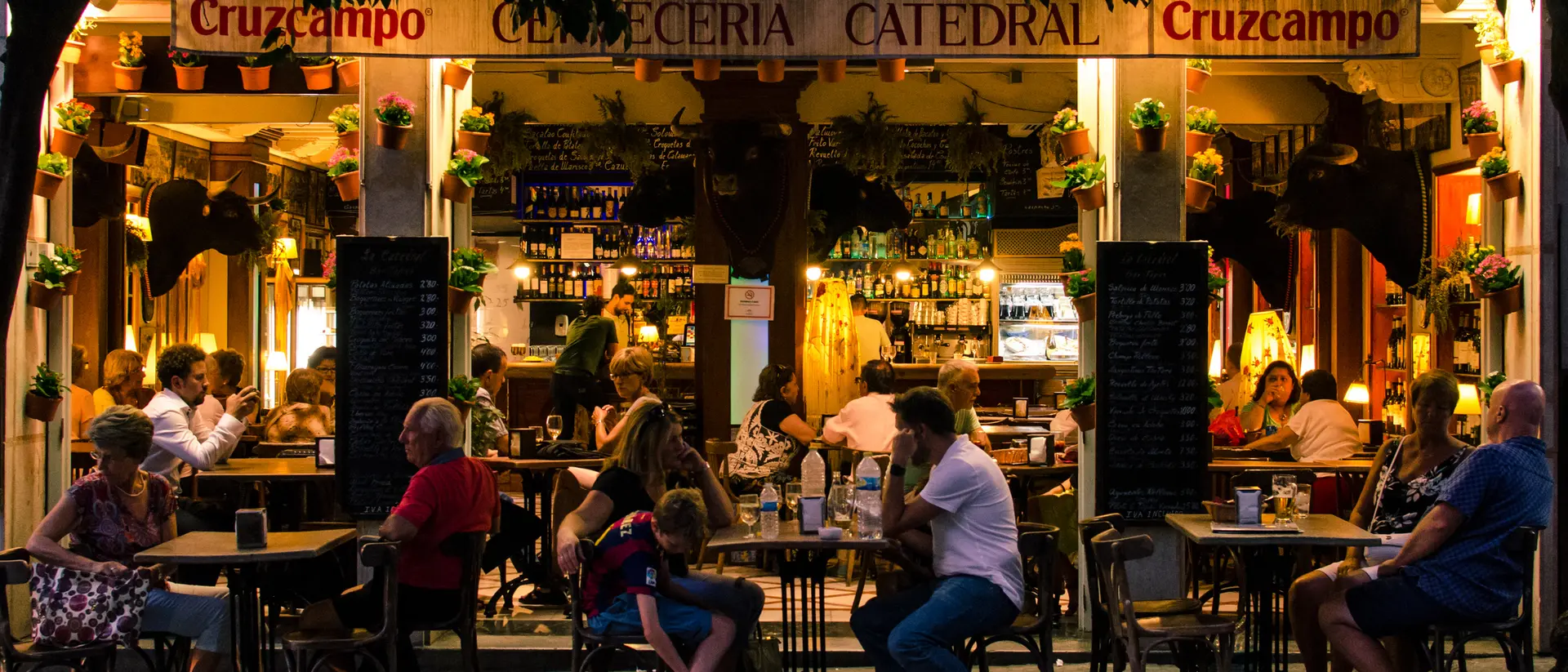 A lively outdoor café with people dining at tables, illuminated by warm lights, surrounded by greenery and a welcoming atmosphere.