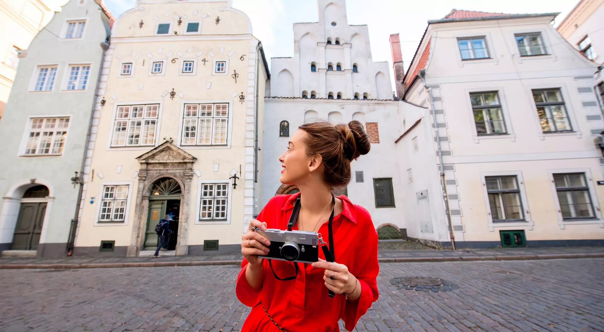 A woman in a red dress holds a camera, smiling as she stands on a cobblestone street with historic buildings in the background.