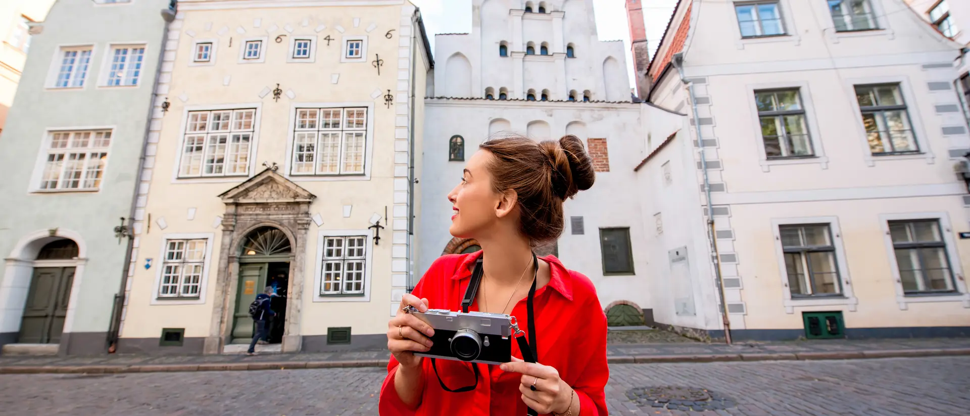 A woman in a red dress holds a camera, smiling as she stands on a cobblestone street with historic buildings in the background.