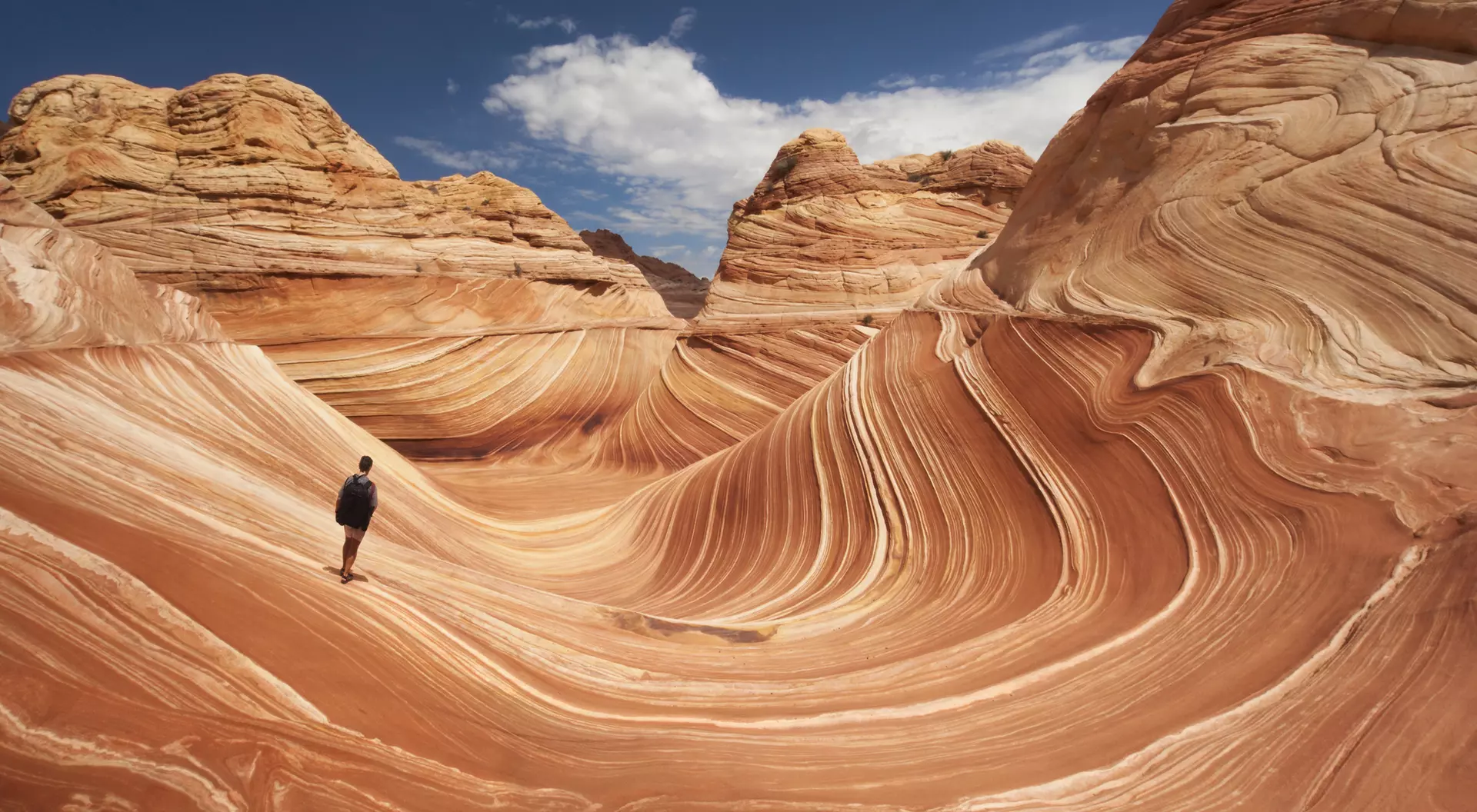 A person stands on colorful, wavy rock formations under a blue sky with scattered clouds.