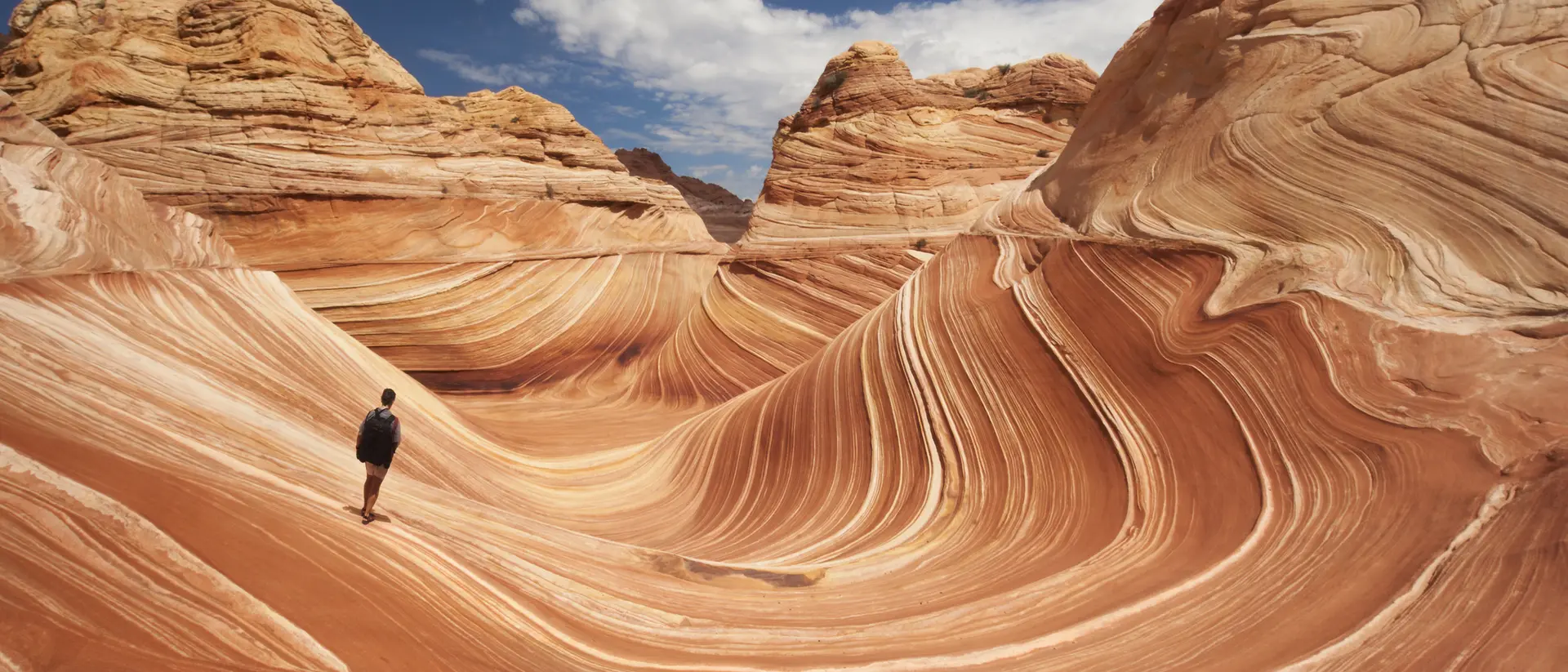 A person stands on colorful, wavy rock formations under a blue sky with scattered clouds.