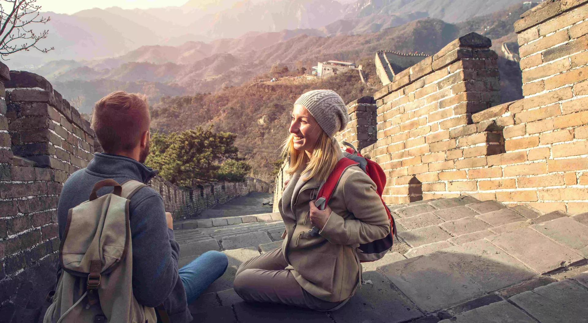 Two hikers sit on the Great Wall of China, enjoying the view of mountains in the background during sunset.