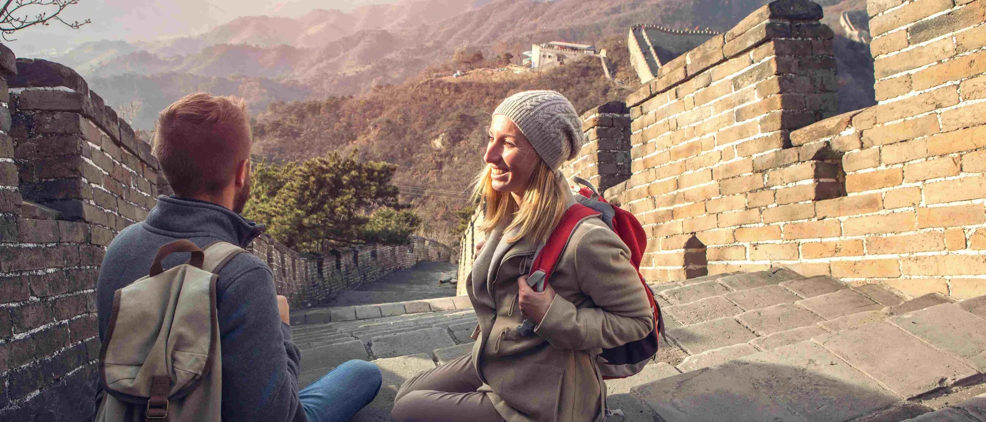 Two hikers sit on the Great Wall of China, enjoying the view of mountains in the background during sunset.