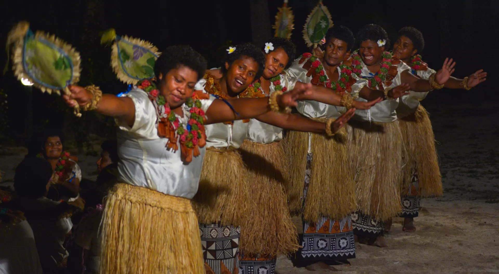 A group of women in traditional attire perform a dance with fans, showcasing vibrant cultural expression under a night sky.