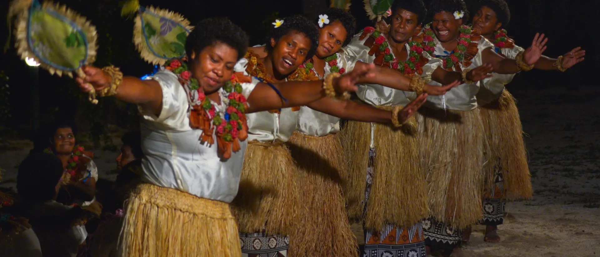 A group of women in traditional attire perform a dance with fans, showcasing vibrant cultural expression under a night sky.