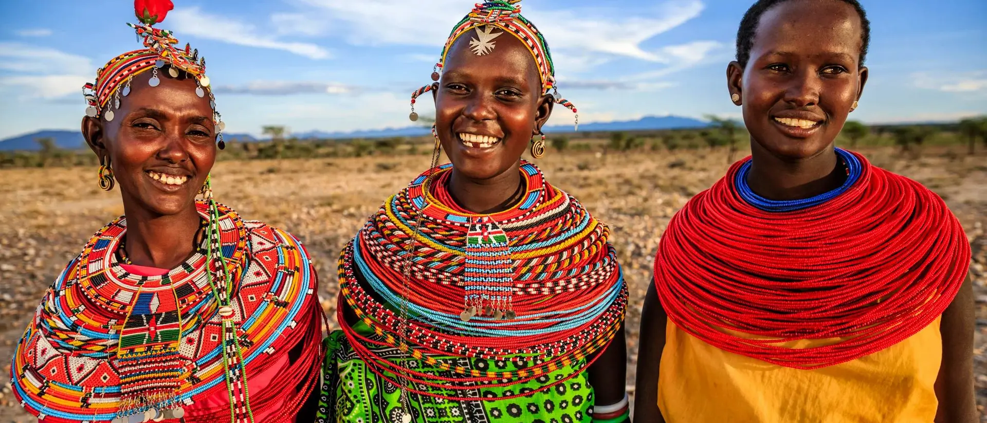 Three smiling women in colorful traditional attire stand together against a scenic landscape.