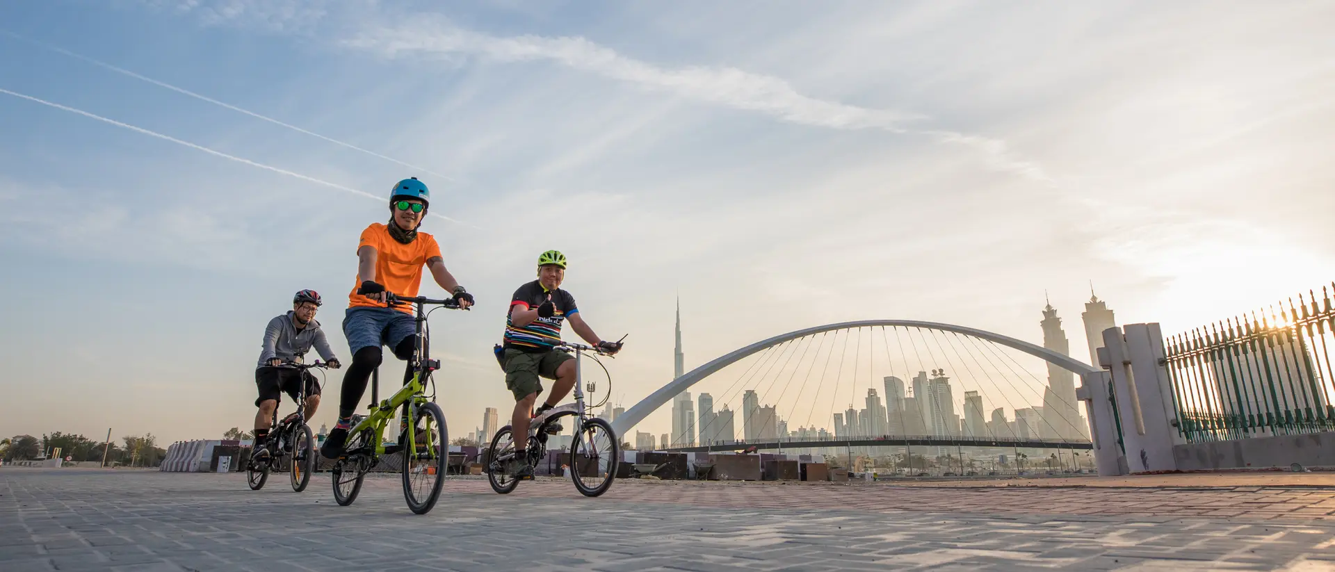 Three children ride bicycles along a path with a city skyline and bridge in the background under a sunset sky.