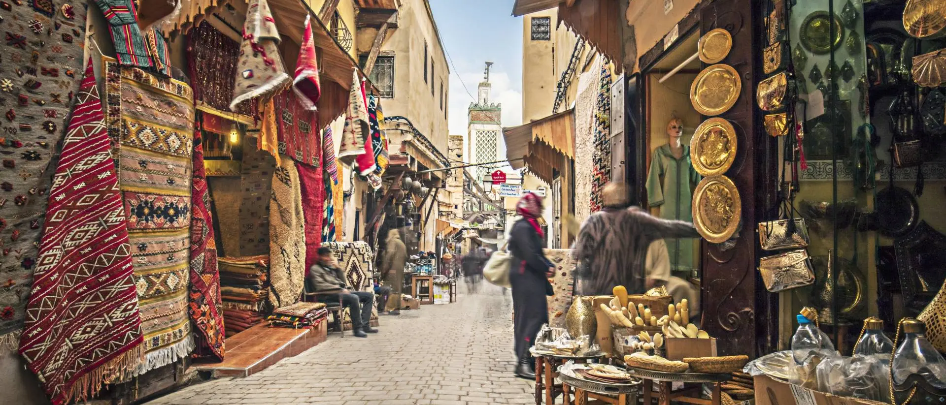 Narrow market street lined with colorful rugs, shops, and people browsing various goods under a clear blue sky.