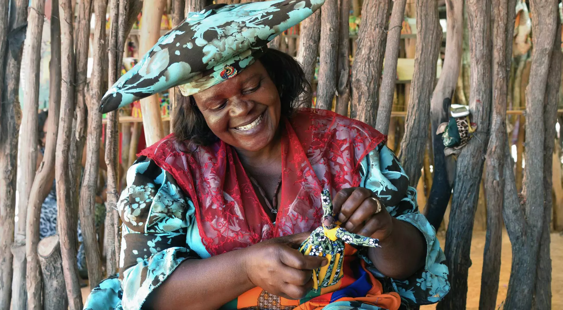 Smiling woman in colorful attire crafts a decorative doll, surrounded by natural wooden elements in a vibrant setting.