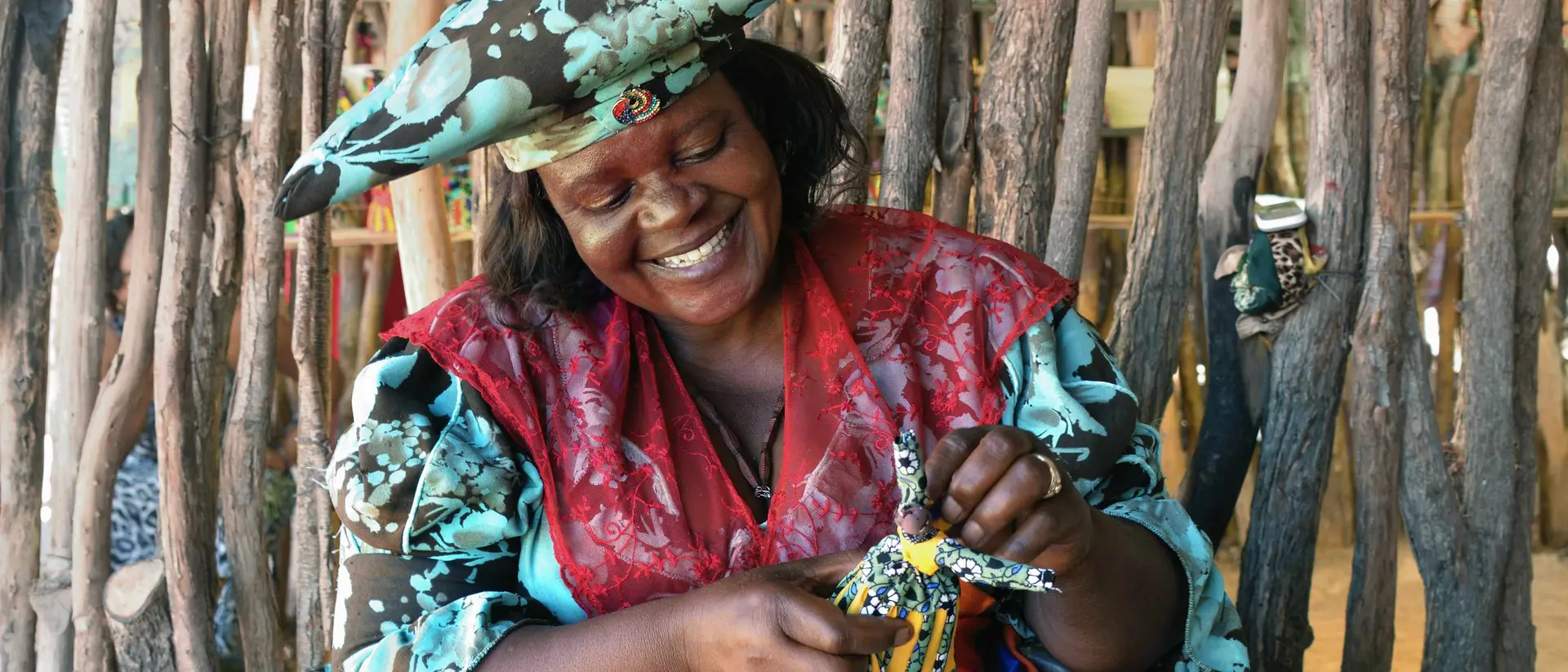 Smiling woman in colorful attire crafts a decorative doll, surrounded by natural wooden elements in a vibrant setting.