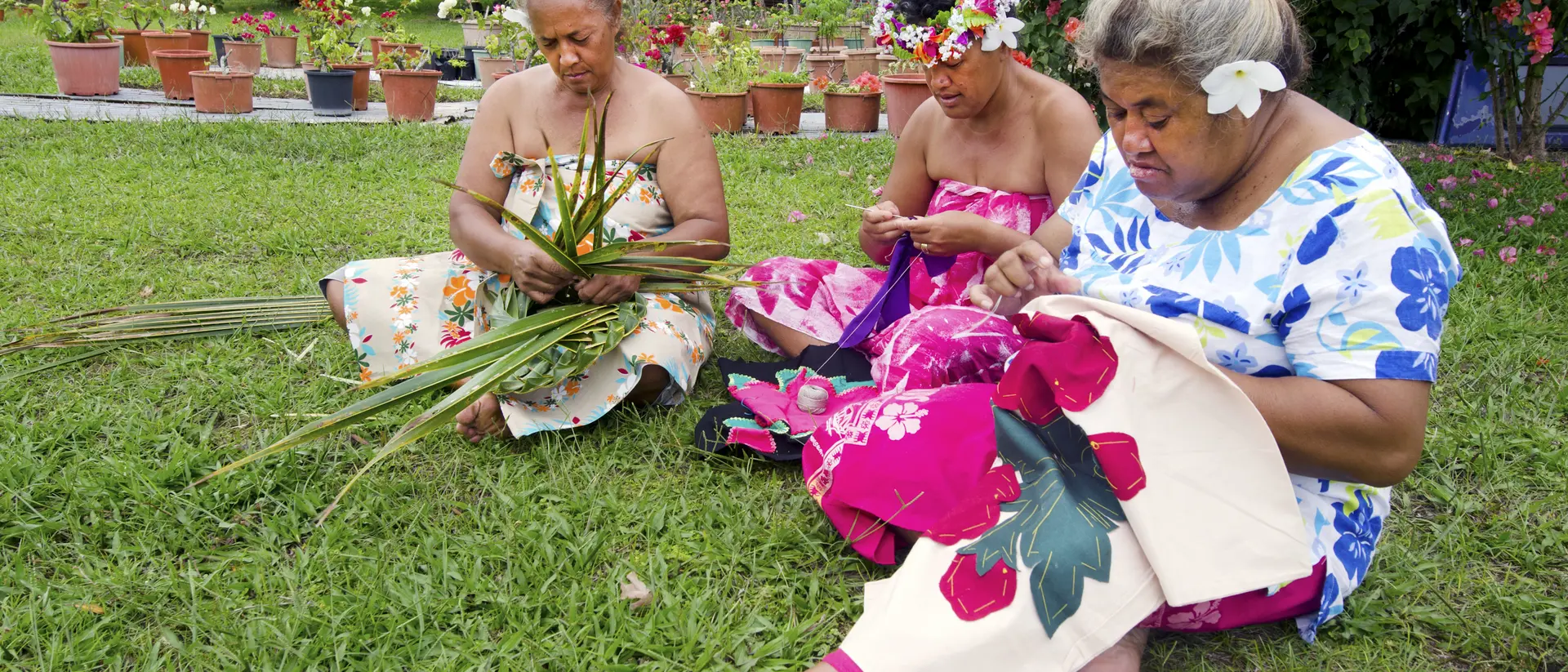 Three women sitting on grass, weaving with leaves and fabric, surrounded by colorful flowers and plants in pots.