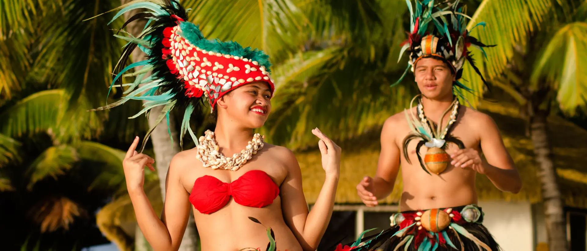 Two dancers in traditional attire perform in a tropical setting, surrounded by palm trees and vibrant greenery.