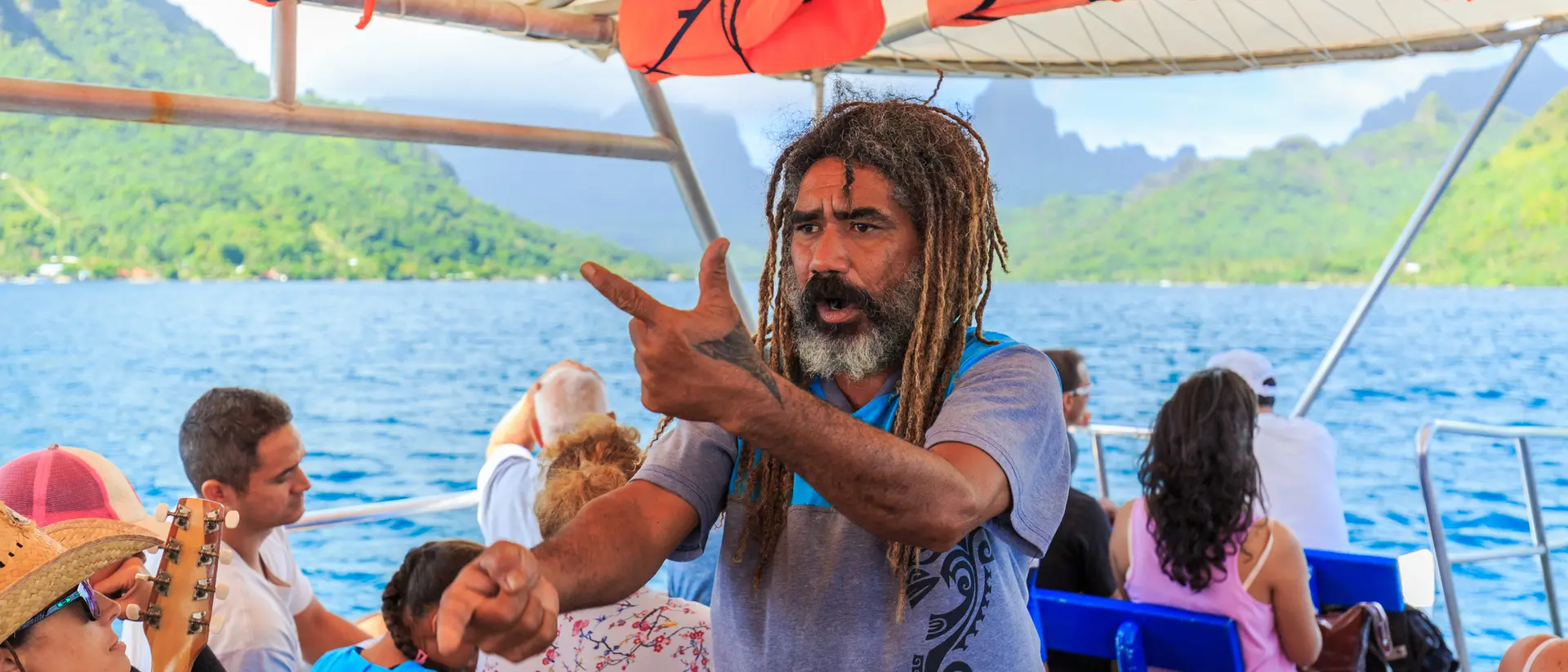 A man with dreadlocks gestures animatedly on a boat, surrounded by people, with scenic mountains and water in the background.