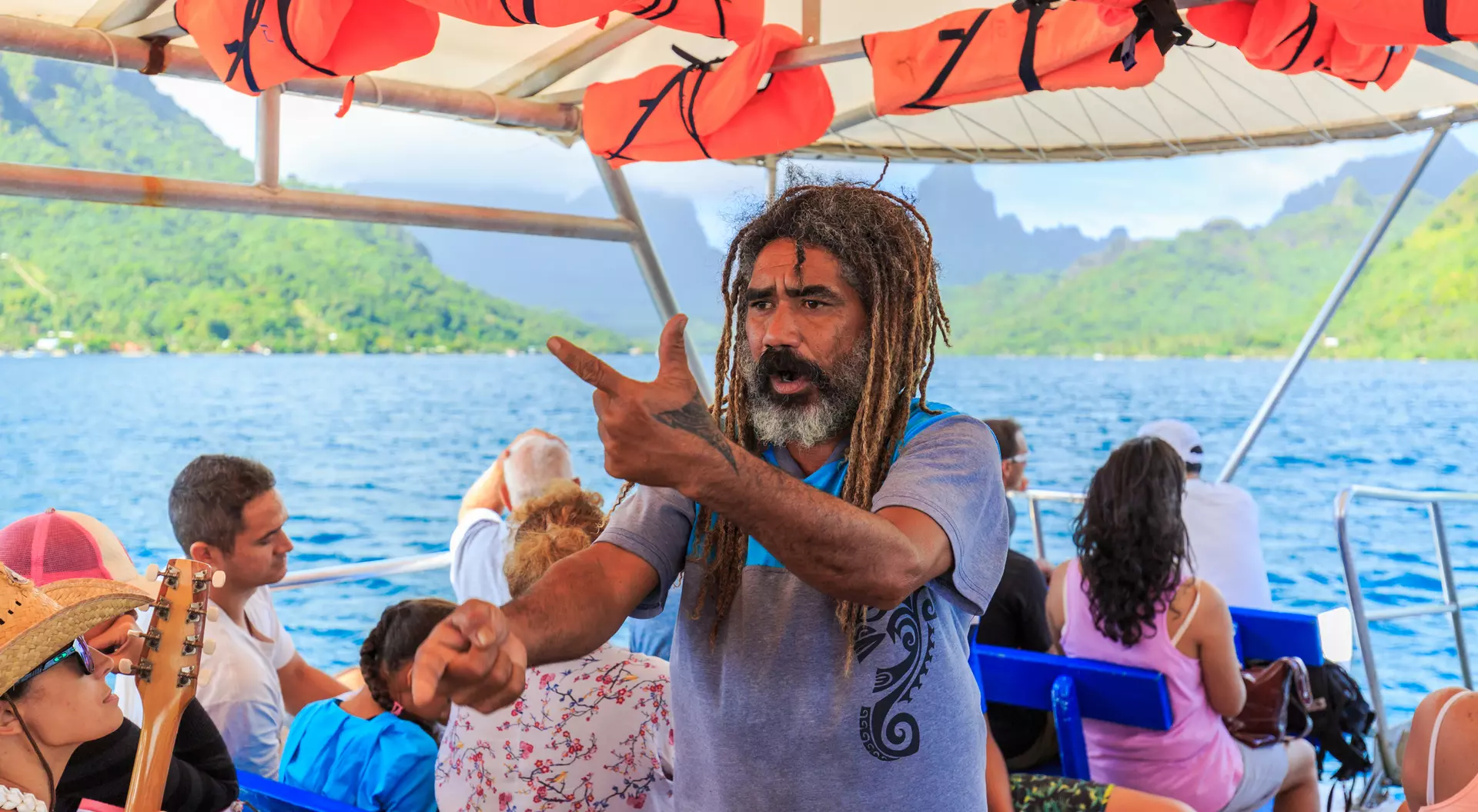 A man with dreadlocks gestures animatedly on a boat, surrounded by people, with scenic mountains and water in the background.