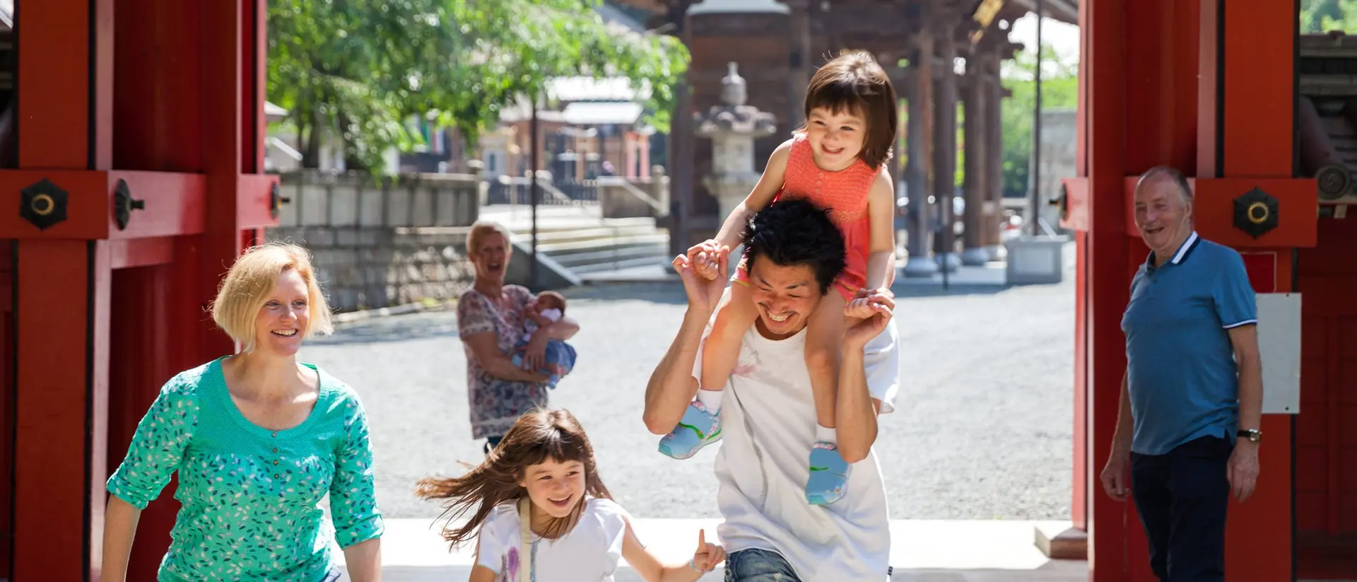 A joyful family walks through a gateway, with a dad carrying a girl on his shoulders and two children running alongside.