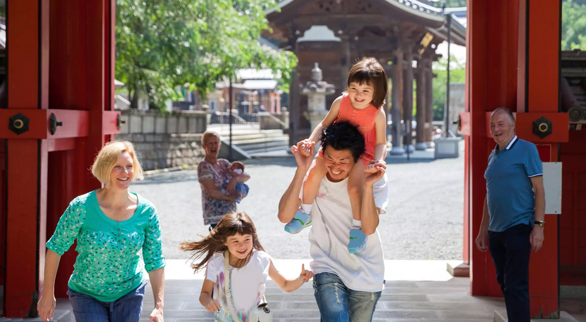 A joyful family walks through a gateway, with a dad carrying a girl on his shoulders and two children running alongside.