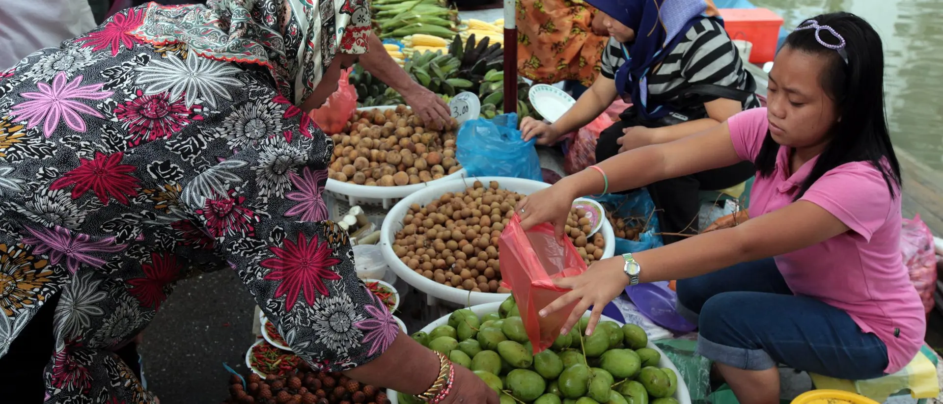 A vibrant market scene with women selling fruits from baskets along a waterway, showcasing various colorful produce.