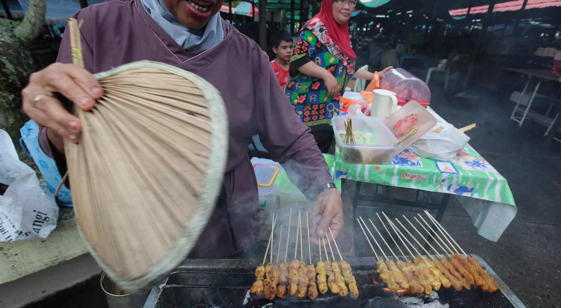 A vendor fans smoky grilled skewers at a market, with people and colorful food stalls in the background.