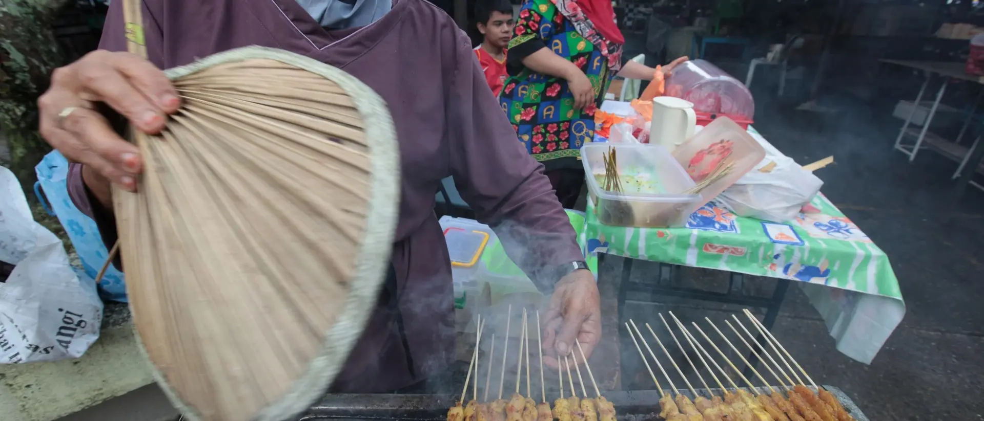 A vendor fans smoky grilled skewers at a market, with people and colorful food stalls in the background.