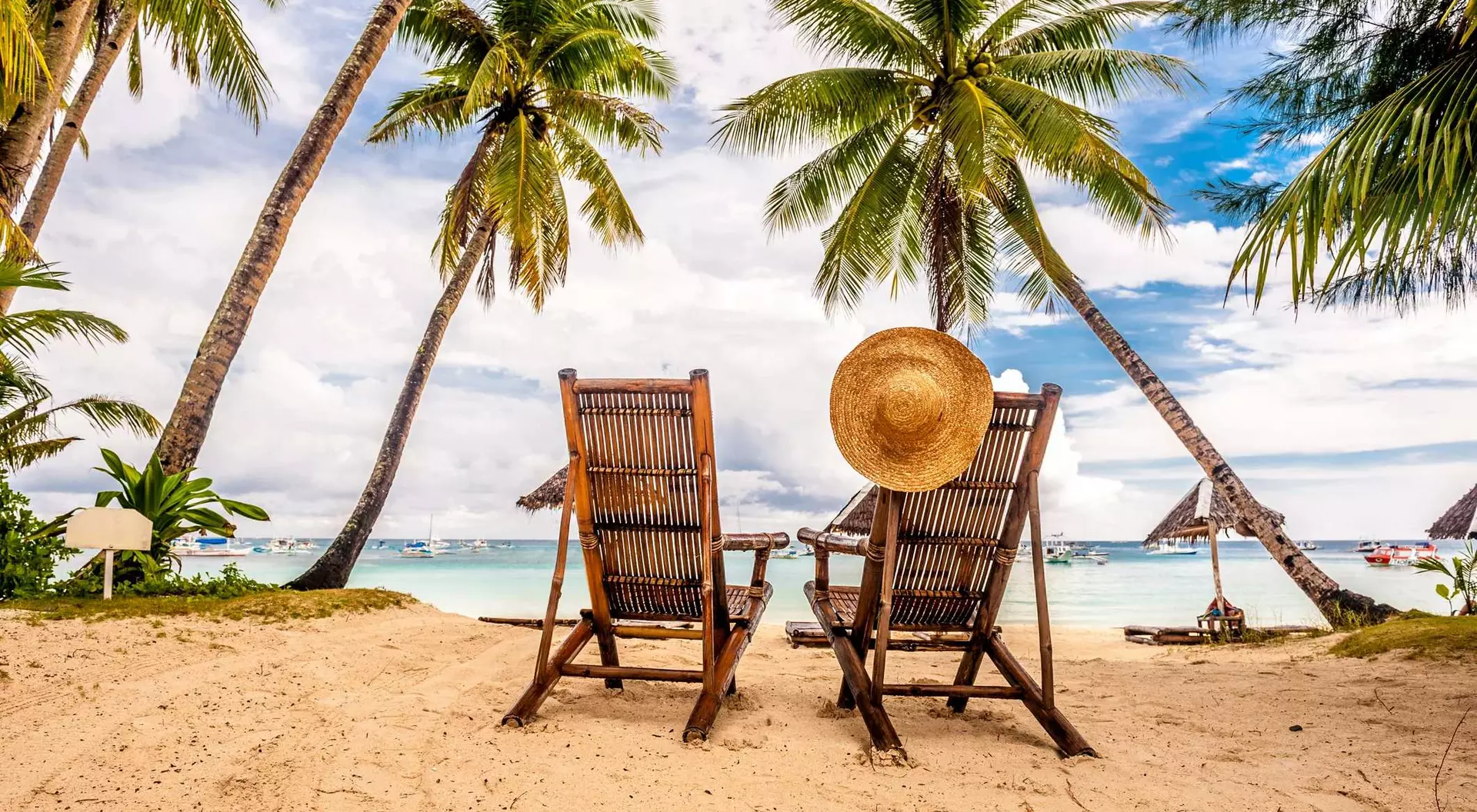Two wooden lounge chairs on a sandy beach, framed by palm trees, with a straw hat resting on the back of one chair.