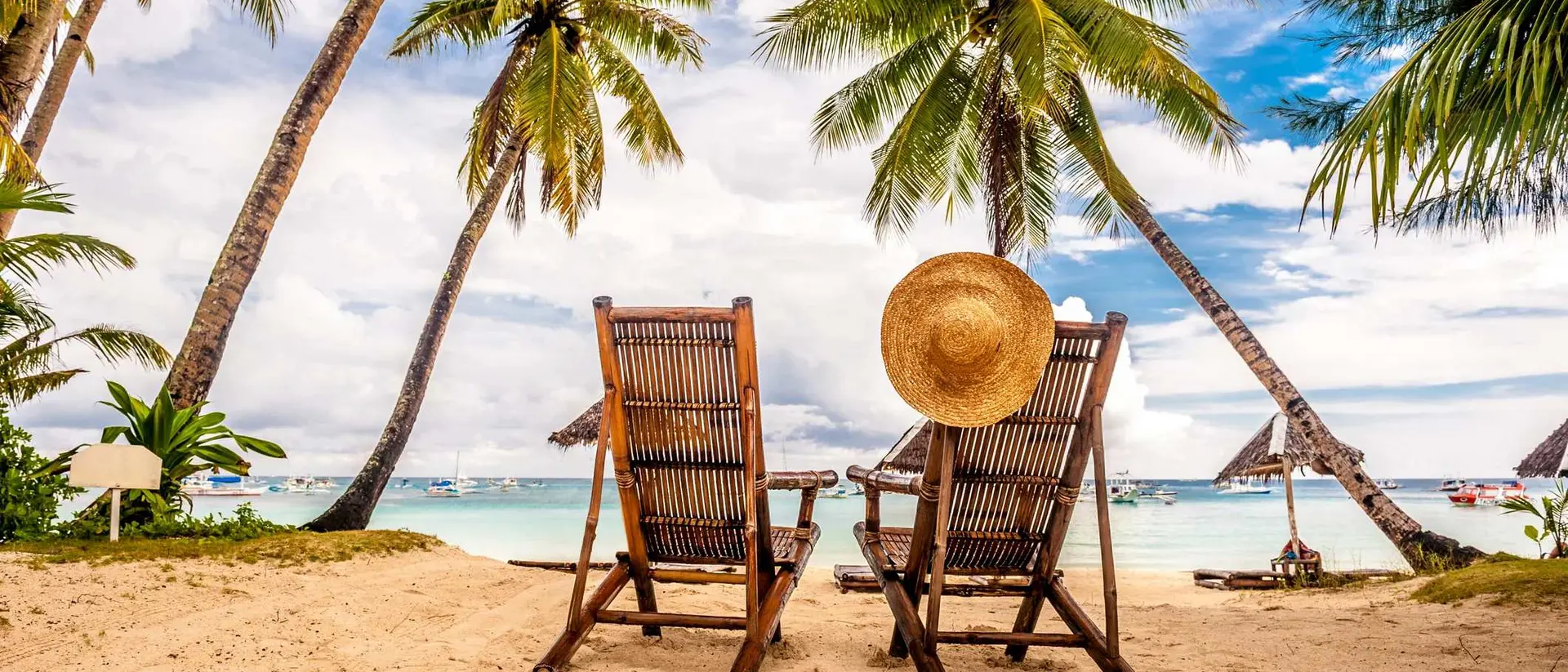 Two wooden lounge chairs on a sandy beach, framed by palm trees, with a straw hat resting on the back of one chair.