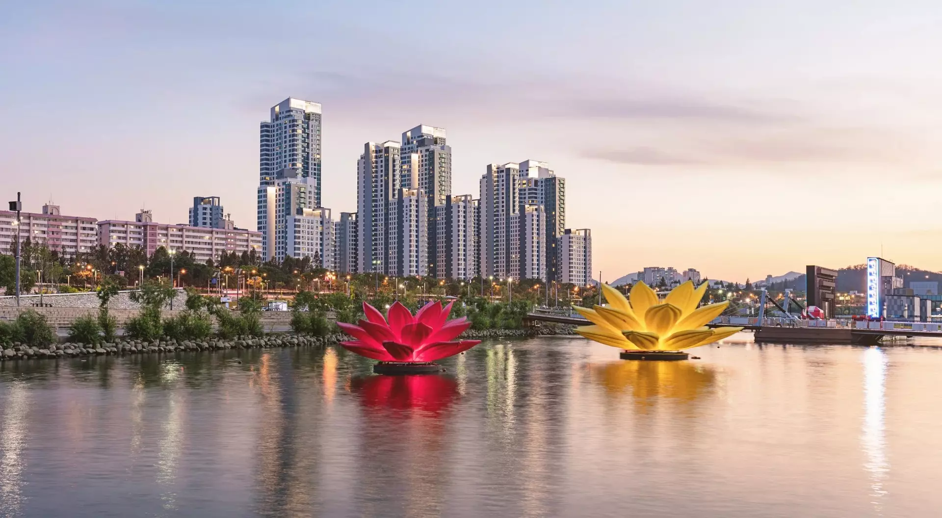 Two large, colorful lotus flower floats in a calm river, with city skyscrapers and a sunset in the background.
