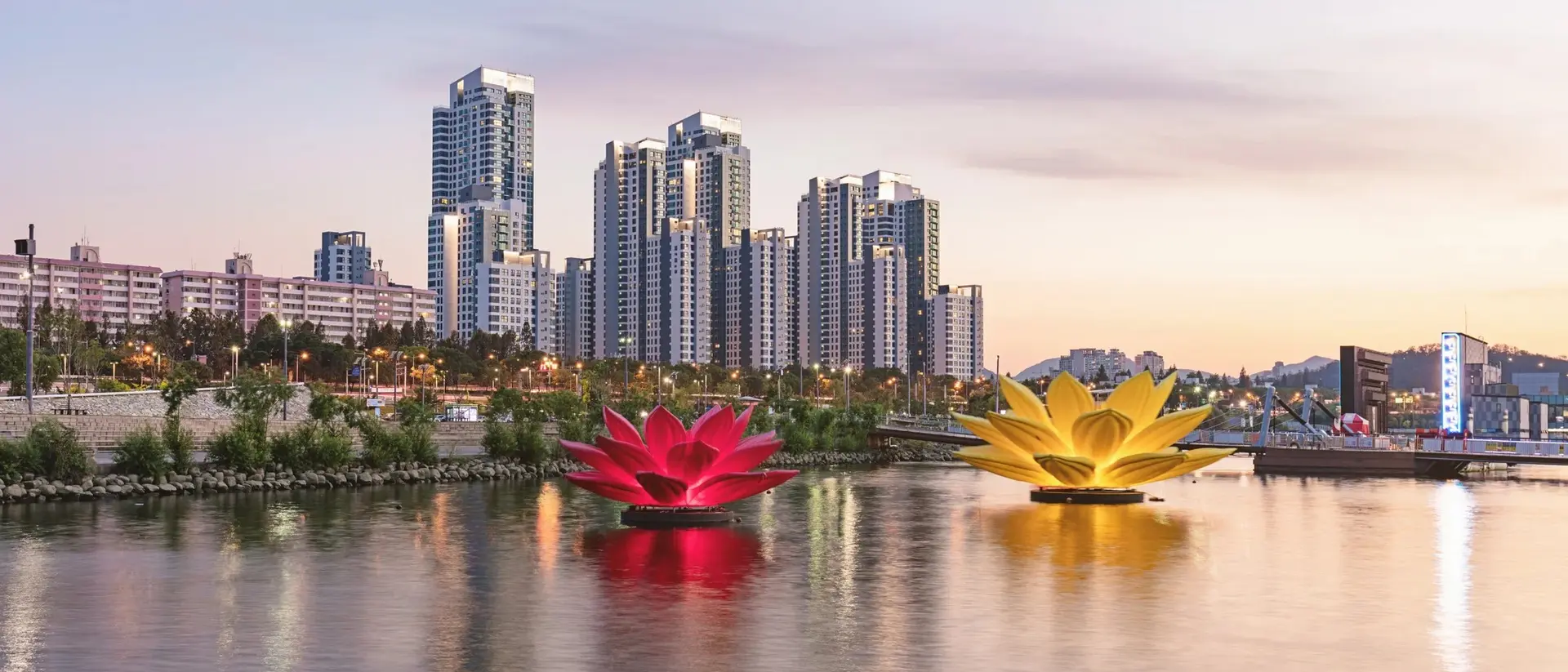 Two large, colorful lotus flower floats in a calm river, with city skyscrapers and a sunset in the background.