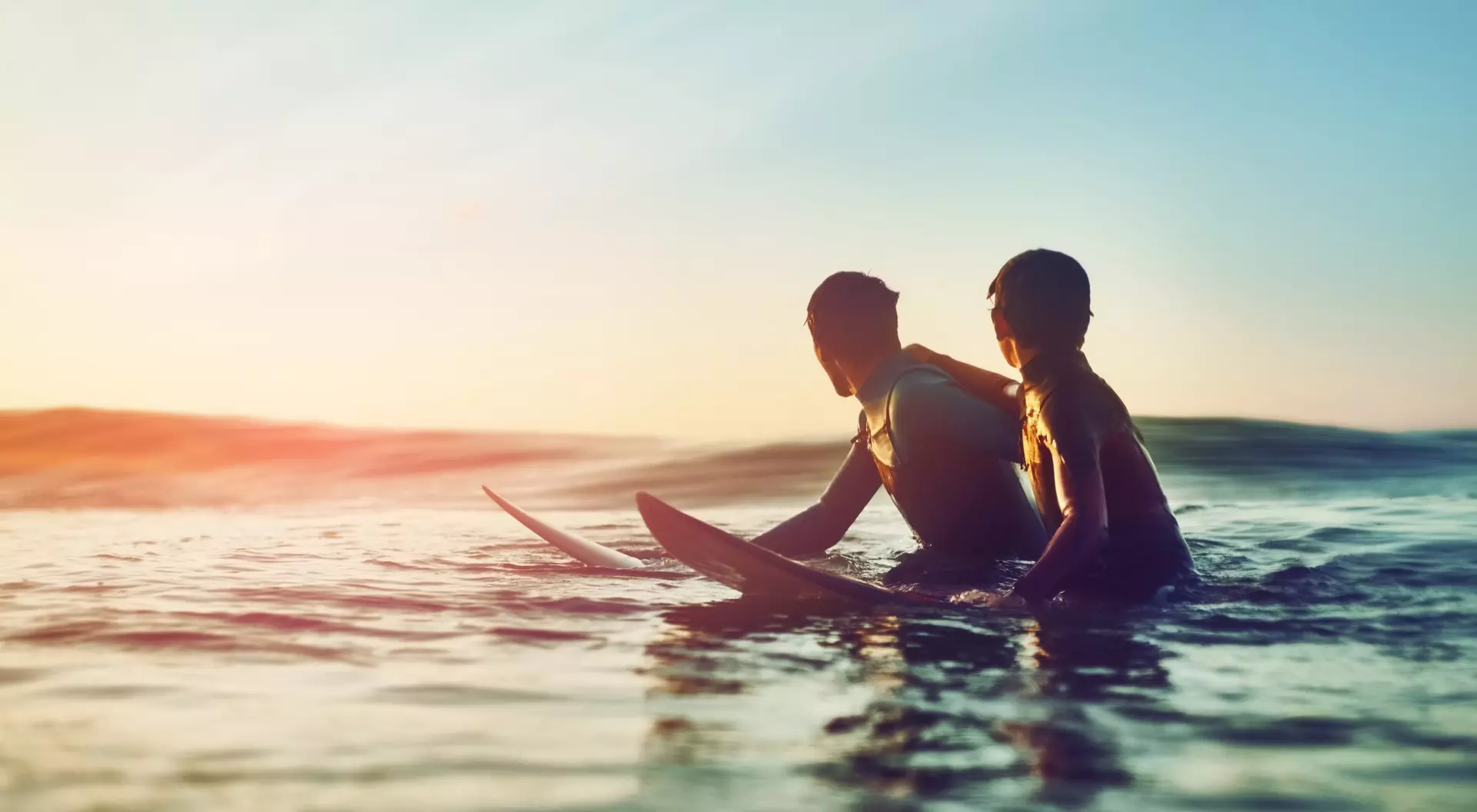 Two surfers sit on their boards in the sea, enjoying a sunset and the calm ocean waves around them.