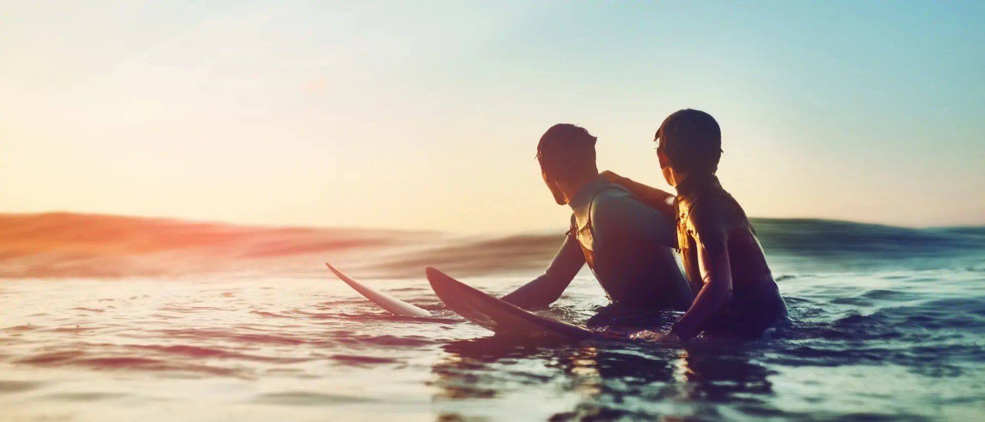 Two surfers sit on their boards in the sea, enjoying a sunset and the calm ocean waves around them.