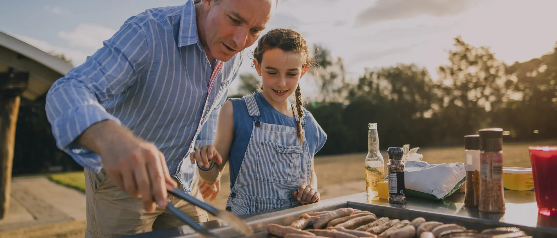 An adult man and a girl are grilling sausages together at a barbecue, enjoying the outdoor setting.