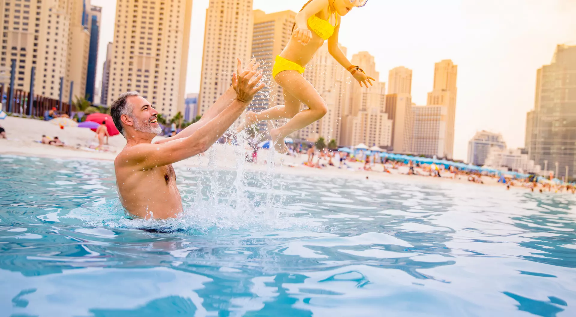 A man lifts a girl in a yellow swimsuit as she jumps playfully into a pool, with city buildings in the background.