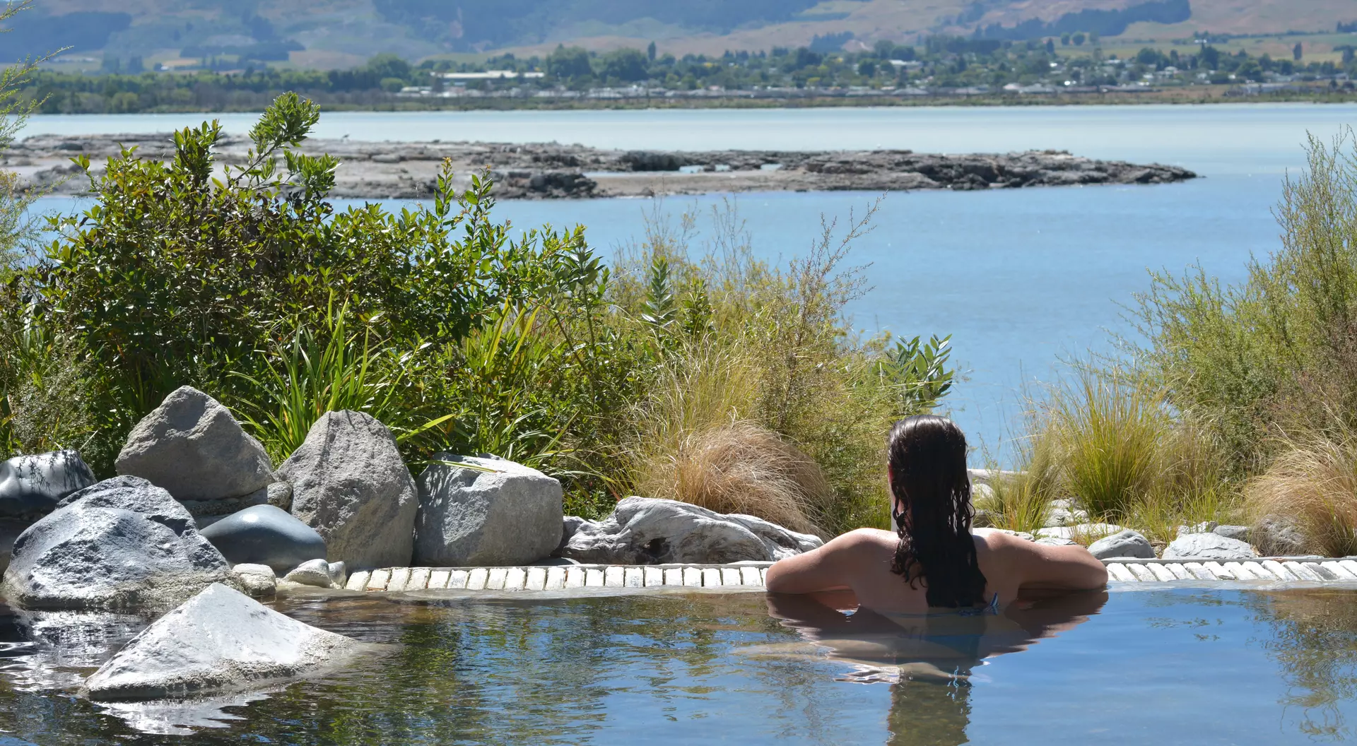 A person relaxes in a hot spring, surrounded by rocks and greenery, with a serene lake and mountains in the background.